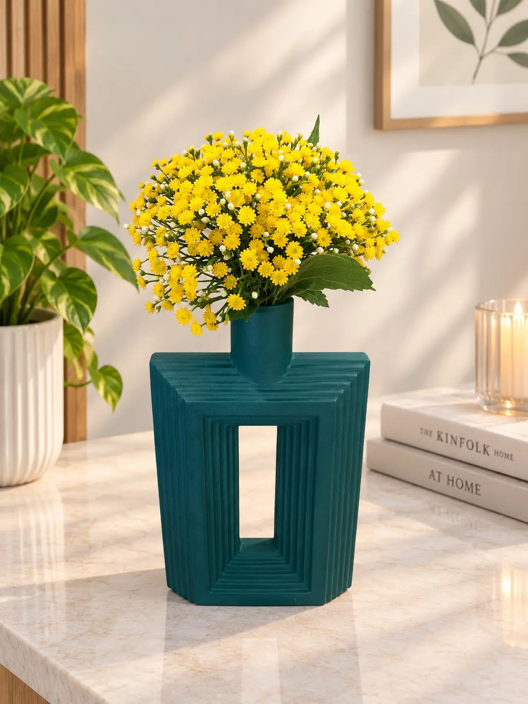 A teal geometric ceramic vase with a hollow center, filled with a bouquet of bright yellow daisy-like flowers, set on a light marble countertop in a styled interior scene.