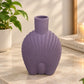 A lavender purple shell-shaped ceramic vase with a short neck, displayed on a light marble countertop in a bright, modern room next to a potted plant and a book.