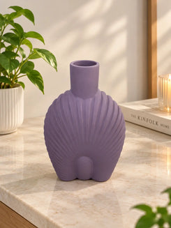 A lavender purple shell-shaped ceramic vase with a short neck, displayed on a light marble countertop in a bright, modern room next to a potted plant and a book.