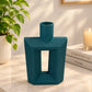 A modern geometric teal ceramic vase with a ribbed texture and a central cutout, displayed on a marble tabletop in a styled home interior next to a potted plant and books.