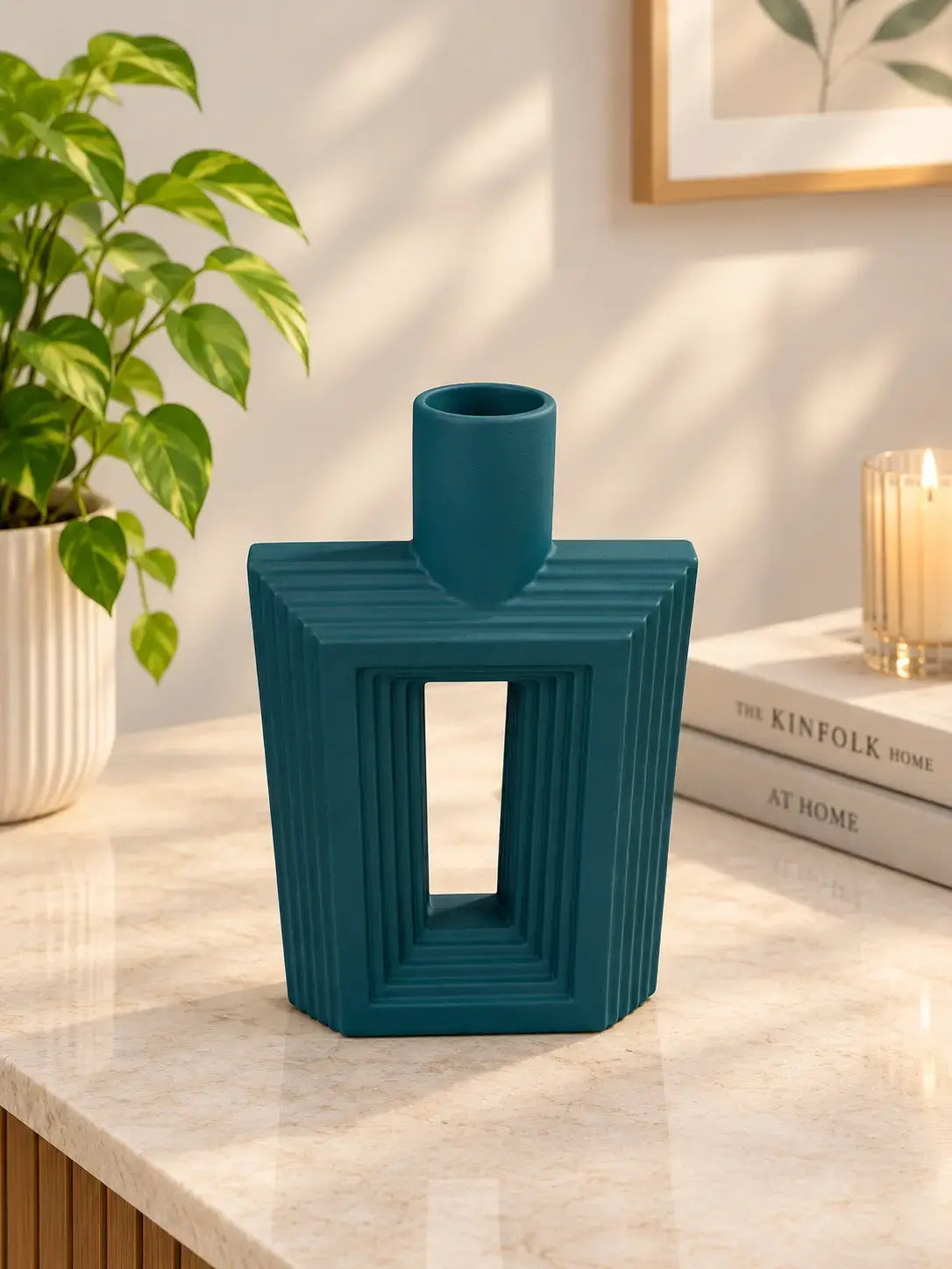 A modern geometric teal ceramic vase with a ribbed texture and a central cutout, displayed on a marble tabletop in a styled home interior next to a potted plant and books.