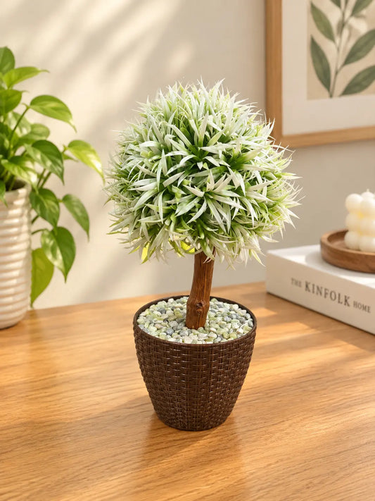 An artificial faux tree topiary with white and green variegated leaves in a brown wicker pot filled with decorative pebbles, displayed on a wooden table in a styled home interior.