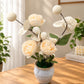 An artificial floral arrangement featuring cream-colored roses, white blossoms, and green leaves in a textured light blue-gray ceramic pot, displayed on a wooden table in a bright, modern room.