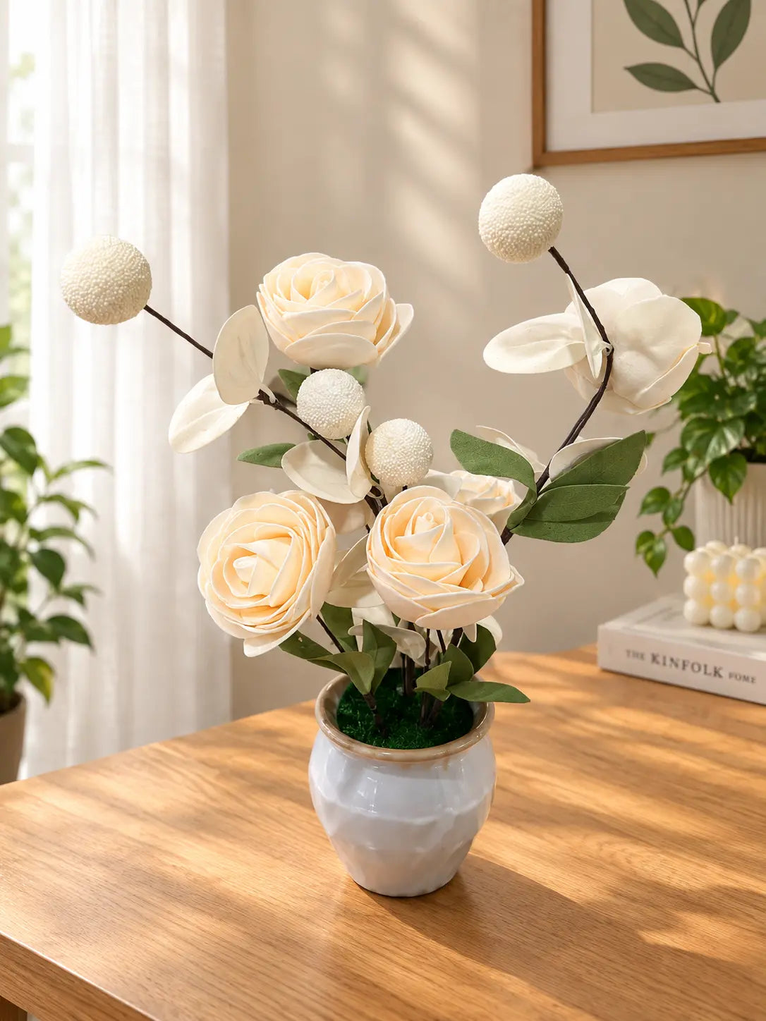 An artificial floral arrangement featuring cream-colored roses, white blossoms, and green leaves in a textured light blue-gray ceramic pot, displayed on a wooden table in a bright, modern room.